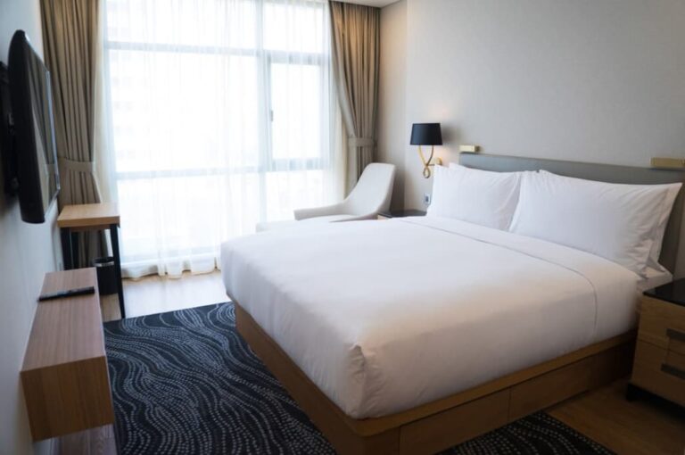 Modern hotel room with a large bed, wall-mounted TV, and sheer curtains over a window